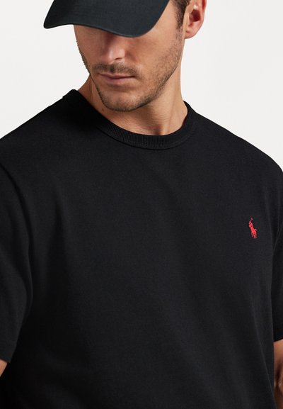 Μαύρο βαμβακερό t-shirt με στρογγυλή λαιμόκοψη και κοντά μανίκια, διακοσμημένο με ένα μικρό κόκκινο κεντημένο λογότυπο στο στήθος. Λείο υλικό.