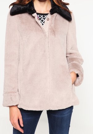 Femme portant une veste en fausse fourrure beige avec un col en fourrure noire, une main dans la poche, associée à un jean bleu foncé et un haut à motifs.