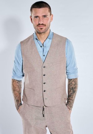 Mann mit kurzen Haaren und Tattoos trägt eine beige Weste und Hose über einem hellblauen hochgekrempelten Hemd, steht mit den Händen in den Taschen.