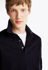 Polo shirt blu navy con colletto strutturato e patta con tre bottoni. Presenta una trama texturizzata e sottili accenti rossi sul colletto.