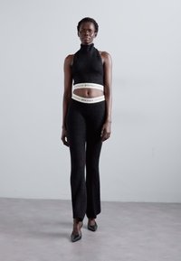Black, textured crop top with high neck paired with high-waisted, flared pants; features branding on the elastic waistband. Pointed heels.