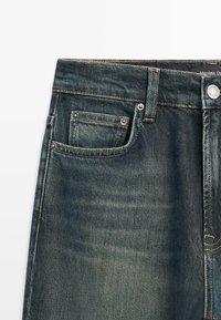 Jeans in denim blu scuro con tasche frontali visibili, cuciture, passanti per cintura e chiusura con bottone metallico su sfondo bianco.