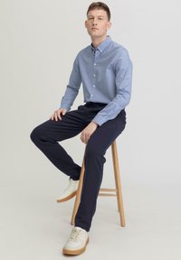 Lichtblauw overhemddasje, marineblauwe broek, witte sneakers met rubberen zolen. Model zit op een houten kruk tegen een effen achtergrond.