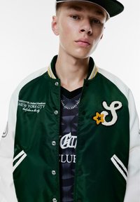 Groene en witte varsityjack met een drukknoopsluiting, geborduurd logo en gestreepte kraag. Gekocht over een zwart gemarmerd netshirt.