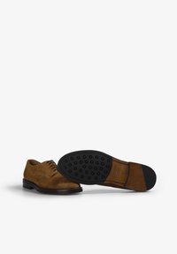 Zapatos de ante marrón con cordones, de exterior suave, punta redondeada y suela de goma con puntiagudas redondeadas para mayor agarre. Costura detallada visible.