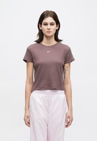 Νεαρή γυναίκα με ίσια καφέ μαλλιά, φορώντας ένα μωβ κοντό μπλουζάκι Nike και ανοιχτό ροζ σατέν αθλητικά παντελόνια, στέκεται μπροστά σε λευκό φόντο.