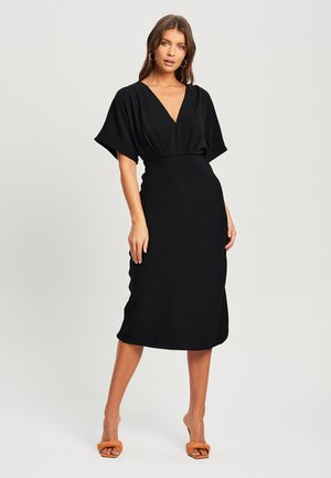 Black dress with a deep V-neck, short sleeves, and fitted silhouette. Fabric appears smooth with a slight sheen. Complemented by tan heels.