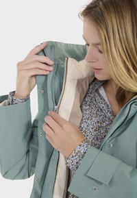 Veste imperméable bleu-vert clair avec fermeture éclair et boutons-pression, dotée d'une doublure beige et d'une chemise à motif floral en dessous.