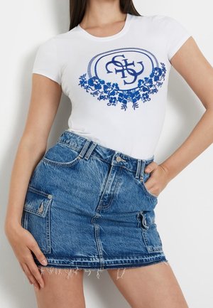 Femme portant un t-shirt blanc ajusté avec un emblème floral bleu et une mini-jupe en jean bleu avec des poches avant et un ourlet effiloché.