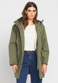 Parka olive avec fermeture éclair à l'avant, accents en boutons et une capuche. L'intérieur possède une doublure douce et texturée. Porté par-dessus un haut blanc.