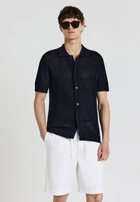 Navyblaues Kurzarmhemd mit einem Strukturstrickmuster, Knopfverschluss vorne und kontrastierendem Kragen. Kombiniert mit weißen Shorts.
