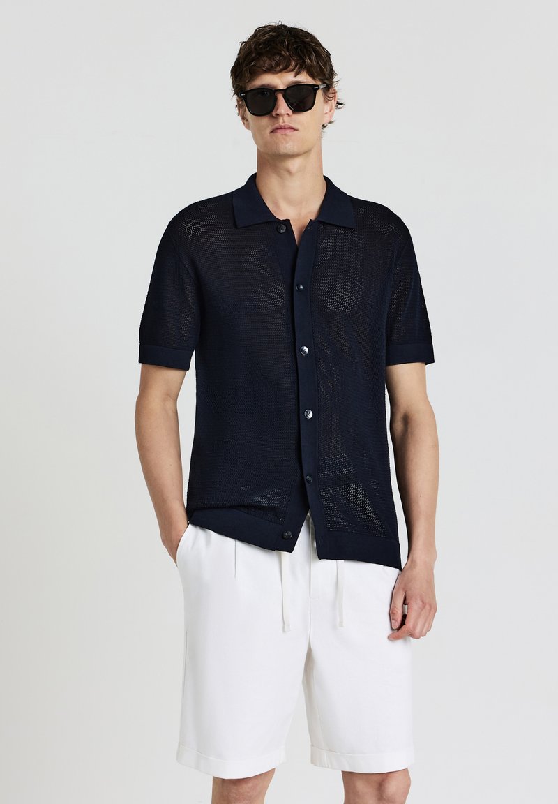 Navyblaues Kurzarmhemd mit einem Strukturstrickmuster, Knopfverschluss vorne und kontrastierendem Kragen. Kombiniert mit weißen Shorts.