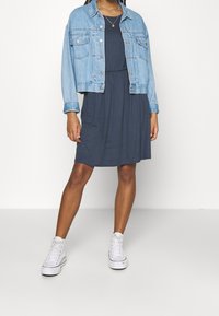 Jeansjacke über einem dunkelgrauen Kleid mit lockerer Passform. Weiße High-Top-Sneaker vervollständigen das Outfit und betonen einen lässigen Look.