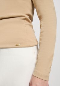 Langärmliges, hellbeiges Oberteil aus einem glatten Stoff, das über eine dezente Textur und ein kleines goldfarbenes Logo-Detail am Saum verfügt.