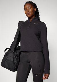 Canottiera nera con zip, maniche lunghe e logo, abbinata a leggings neri. La modella tiene una borsa da palestra nera. Design semplice e funzionale.