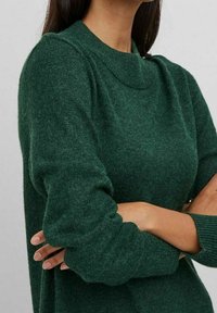 Maglione lavorato a maglia verde scuro con scollo rotondo e polsini a coste. Il tessuto appare morbido e con una texture, con una vestibilità leggermente ampia.