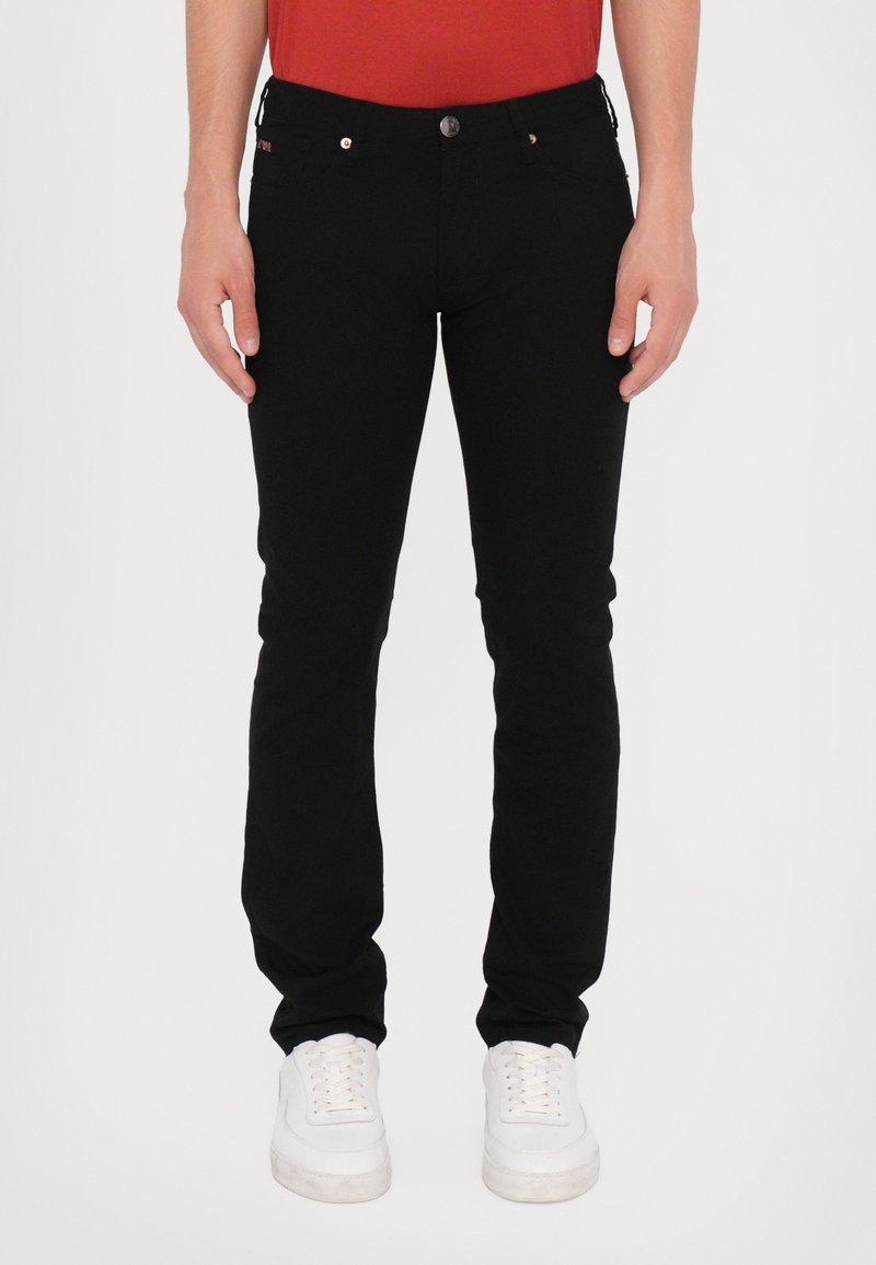 Schwarze Slim-Fit-Jeans mit Knopfverschluss, Fünf-Taschen-Design und subtilen goldenen Akzenten. Tragen mit weißen Sneakers.