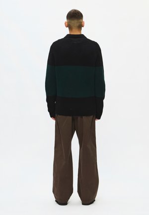 Personne debout de dos portant un pull rayé noir et vert foncé et un pantalon marron ample sur un fond blanc uni.