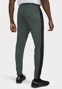 Mannen sportbroeken in donkergroen met zwarte zijstrepen. Kenmerken zijn een taps toelopende pasvorm, elastische tailleband en ritsen aan de voeten. Glad stofoppervlak.