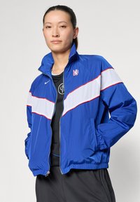 Blå nylon zip-jacka med vita och röda horisontella ränder, hög krage, ribbad linning och logotypdetalj på bröstet.