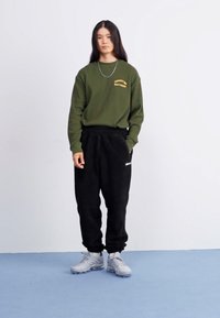 Olivovozelená mikina s žltým textom, kombinovaná s čiernymi fleece nohavicami. Model má na sebe šedé športové topánky a stojí na svetlomodrej podlahe.