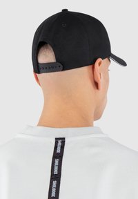 Black cap with a curved peak and snapback closure; worn with a light grey shirt featuring a black vertical logo detail on the back.