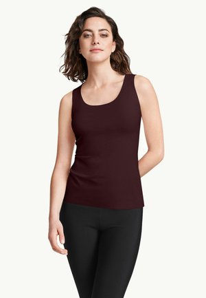 Giovane donna con capelli castani fino alle spalle che indossa un top marrone scuro senza maniche e pantaloni neri, in piedi su uno sfondo semplice.