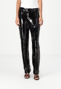 Pantalons noirs en sequins avec une coupe droite, présentant une texture brillante et une taille élastique. Portés avec des sandales noires à lanières.