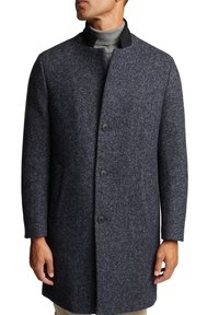 Uomo che indossa un cappotto in lana a spina di pesce grigio scuro con colletto nero sopra un maglione a collo alto grigio chiaro.