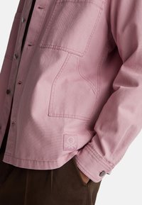 Różowa kurtka z płótna z czterema kieszeniami, zatrzaskami i teksturowanym wykończeniem. Haftowane logo na dolnej lewej kieszeni.
