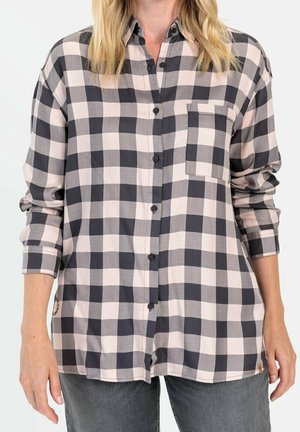 Personne portant une chemise ample à manches longues à carreaux noirs et beiges, avec boutons, poche frontale et manches retroussées.