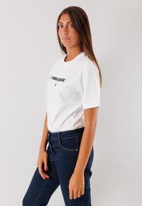 Maglietta in cotone bianco a maniche corte, con testo nero sul fronte. Abbinata a jeans in denim blu scuro. Texture liscia, vestibilità casual.