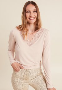 Langärmliges, hellpinkfarbenes Oberteil mit V-Ausschnitt und Spitzenbesatz. Kombiniert mit beigen Schlange-Muster-Hosen in glatter Textur.