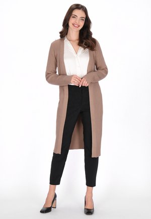 Cardigan long côtelé de couleur marron clair, à ouverture frontale, assorti à un chemisier blanc et un pantalon noir. Des talons hauts noirs complètent la tenue.