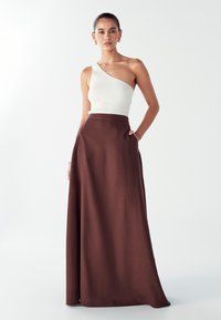 Vrouw in een witte ribbeltop met één schouder en een hooggetailleerde bruine maxirok met zakken, staand tegen een effen witte achtergrond.