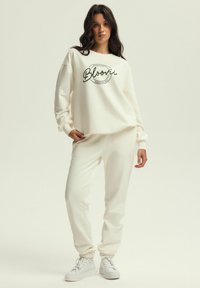 Biała bluza z kapturem z czarnym logo "Bloovii", w zestawie z dopasowanymi białymi dresami. Materiał wydaje się miękki, z rypsowanymi mankietami i luźnym krojem.