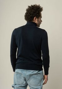 Navyblauwe coltrui met geribde manchetten en zoom, gemaakt van zacht breimateriaal. Model is van achteren te zien, in combinatie met lichte denim jeans.
