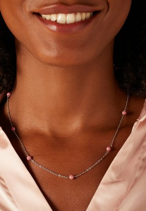 Fijne zilveren ketting met meerdere roze kralen gelijkmatig verdeeld over de ketting, rustend op een gladde huidtint, gecombineerd met een zijden blouse.