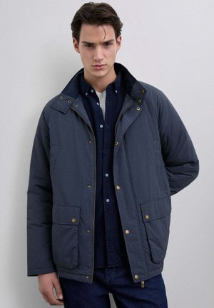 Chaqueta aislante de color azul marino con un alto cuello, herrajes dorados, dos bolsillos frontales y cierre frontal con cremallera sobre una camisa abotonada oscura.