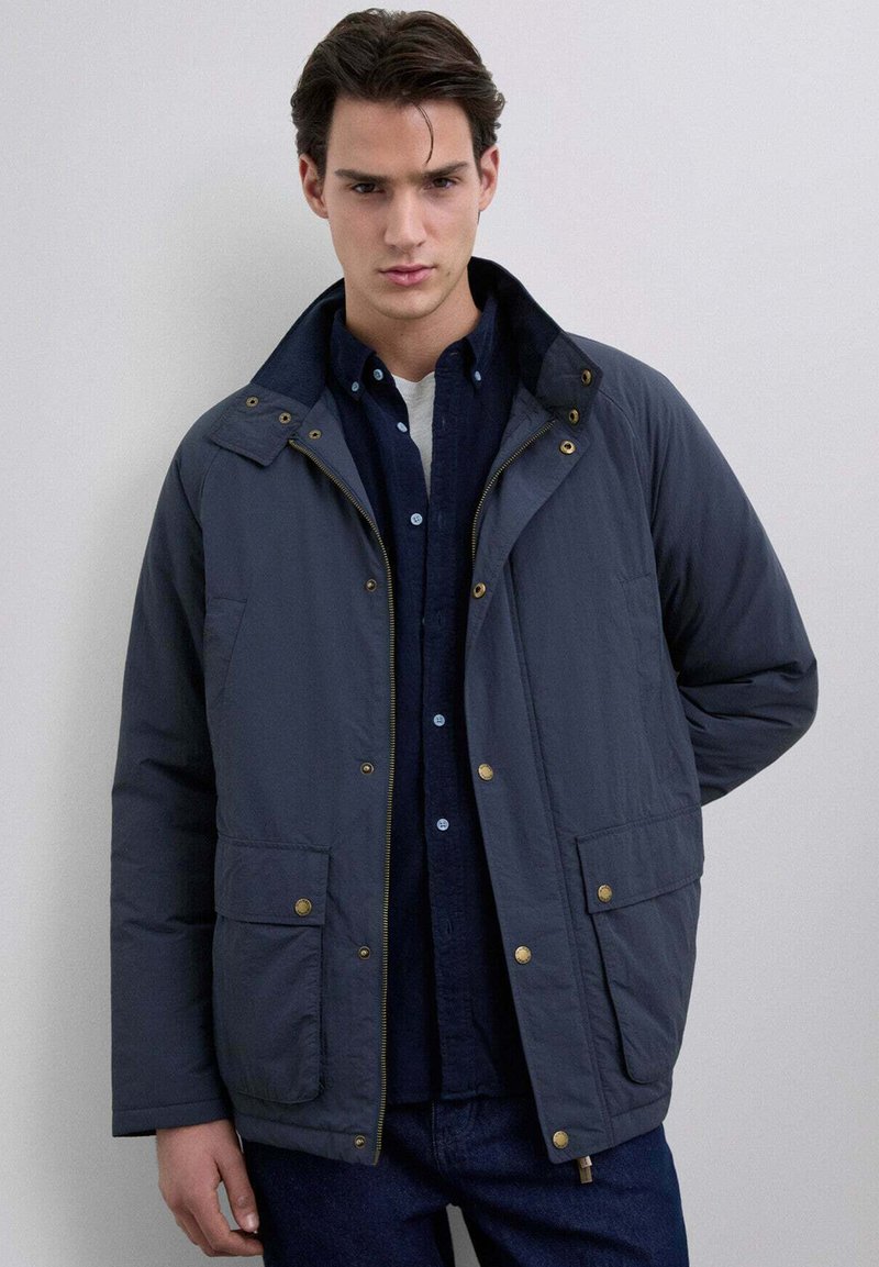 Laineen sininen eristetty takki, jossa on korkea kaulus, kultaiset yksityiskohdat, kaksi etutaskua ja edestä suljettava vetoketju tumman nappikauluspaitan päällä.