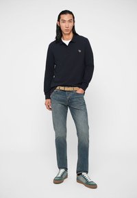 Camisa polo de manga longa azul-marinho com um logo, jeans azul claro e ténis verdes e brancos, combinados com um cinto bege, em pé contra um fundo simples.