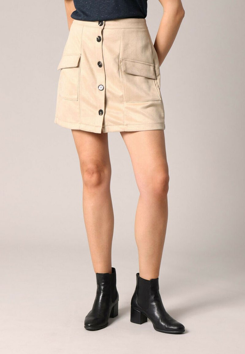 Jupe en velours côtelé beige de coupe évasée, avec fermeture à boutons sur le devant, deux grandes poches plaquées et une texture douce. Portée avec des bottines noires.