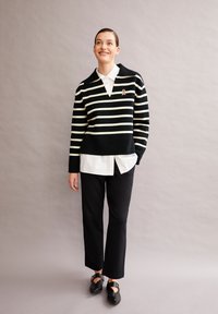 Zwart-wit gestreepte trui met een brede kraag, gedragen over een witte blouse met kraag. Donkere broek en zwarte schoenen met gespen maken de outfit compleet.