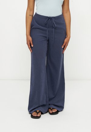 Personne portant un pantalon bleu marine à jambes larges avec taille à cordon, un haut sans manches de couleur claire et des tongs noires, debout devant un fond blanc.