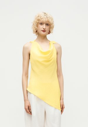 Γυναίκα με μπούκλες ξανθά μαλλιά που φοράει κίτρινο ασύμμετρο αμάνικο τοπ και λευκό παντελόνι σε απλό λευκό φόντο.