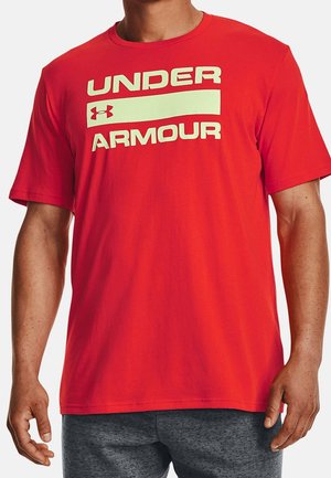 Mann trägt ein rotes T-Shirt mit kurzen Ärmeln mit dem Text "UNDER ARMOUR" und Logo, kombiniert mit dunkelgrauen Hosen.