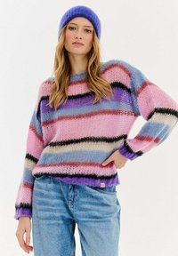 Pletený svetr s horizontálními pruhy v růžové, modré, fialové a černé barvě, s volnějším střihem a žebrovanými manžetami. Nosený přes modré džíny.