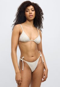 Σετ μπικίνι από ύφασμα ανοιχτού μπεζ χρώματος με υφασμάτινο μοτίβο. Τρίγωνο τοπ με λεπτές τιράντες· το κάτω μέρος διαθέτει ρυθμιζόμενα κορδόνια στα πλαϊνά.