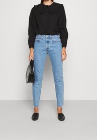 Blouse noire avec un col volant, jeans bleu taille haute, sac à main noir et chaussures noires à enfiler. Denim délavé, légère effilochure à l'ourlet.