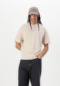 Beige short-sleeve t-shirt with a front graphic, paired with dark jeans and a faded cap. The fabrics appear soft and casual.